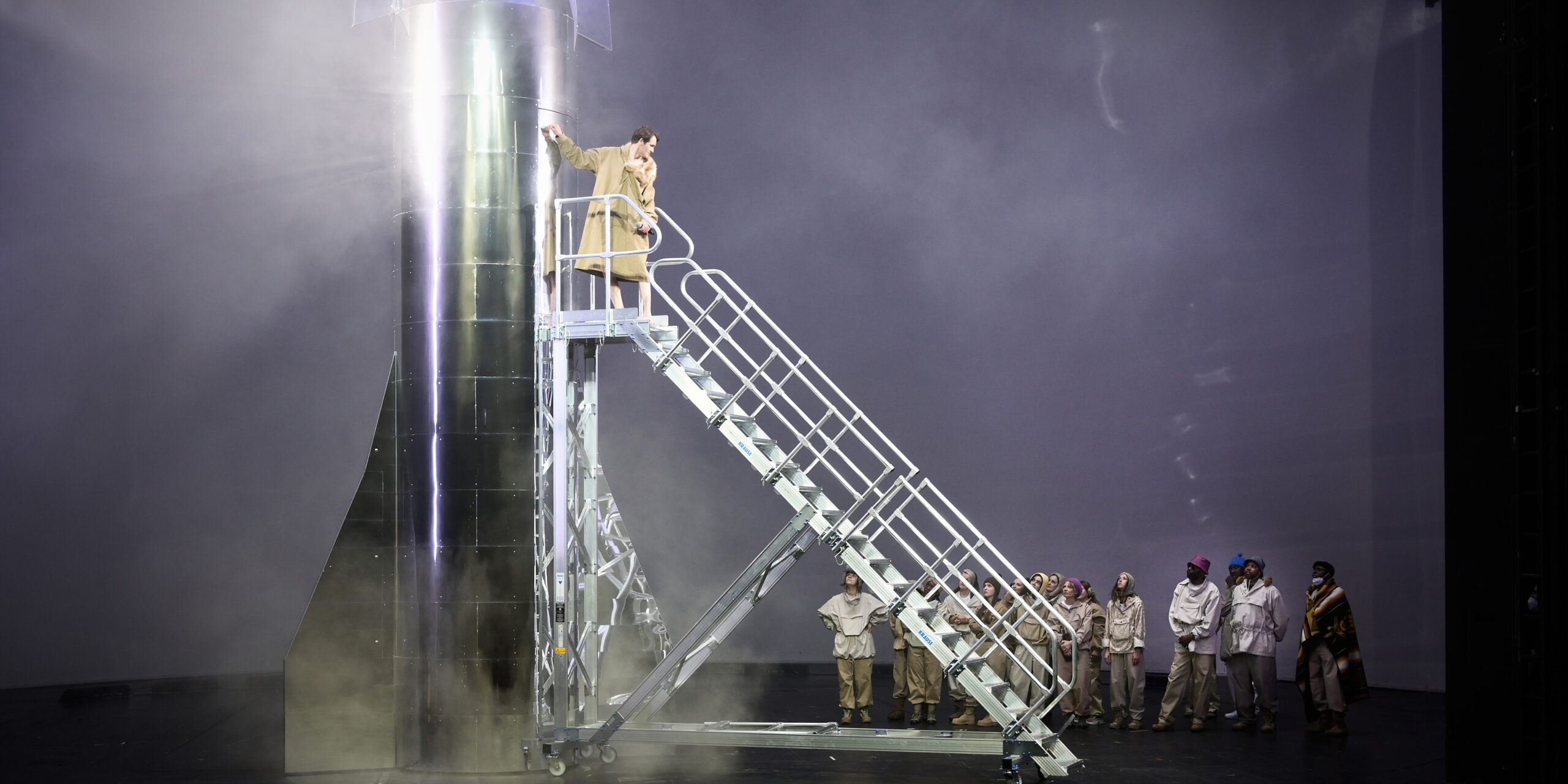 Der Schauspieler Fabian Hinrichs steht auf der obersten Sprosse einer 3 Meter hohen Leiter, die zum Eingang in eine 5 Meter hohe silbern glänzende Rakete führt